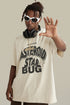  Man in cream graphic tee raises hand forward while wearing white sunglasses and headphones.