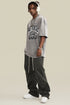 Man in gray tee and dark pants wearing headphones and chains, standing angled to the left.
