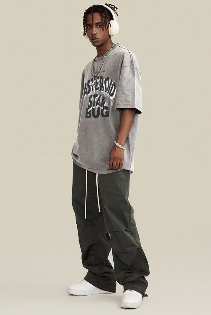 Man in gray tee and dark pants wearing headphones and chains, standing angled to the left.