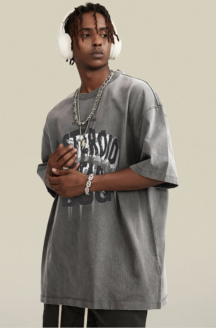 Man in gray tee with headphones and layered jewelry clasping hands in front.