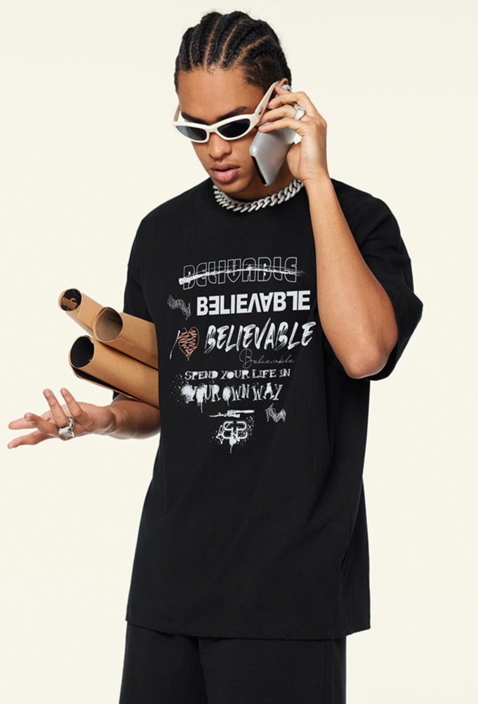Model wearing black graphic tee talking on phone, holding rolled posters under arm.