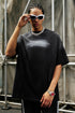 Man in black gradient quote tee adjusts sunglasses while posing against industrial backdrop.