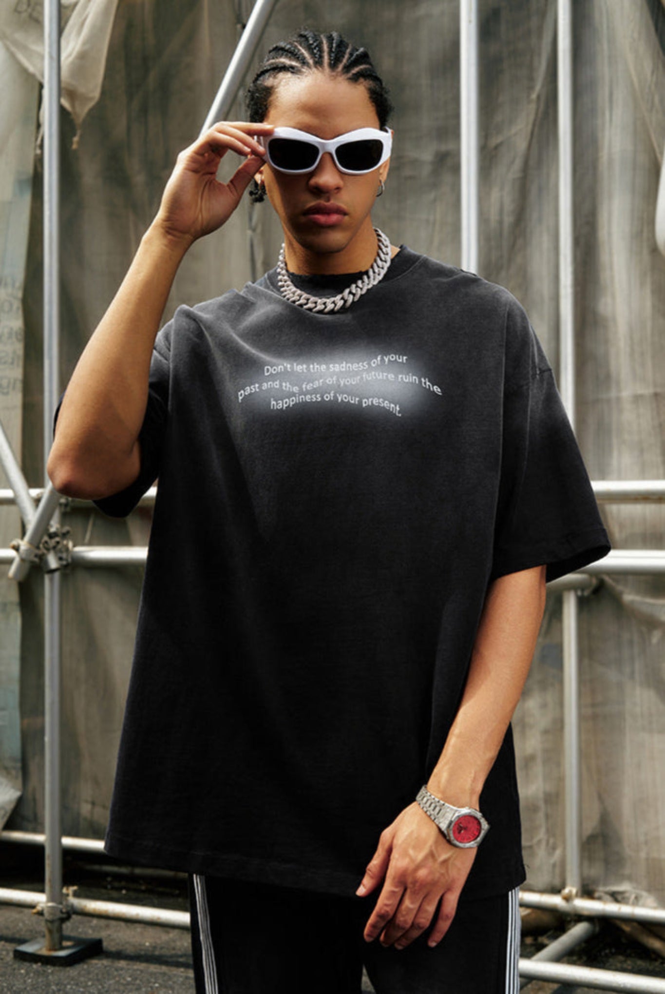 Man in black gradient quote tee adjusts sunglasses while posing against industrial backdrop.