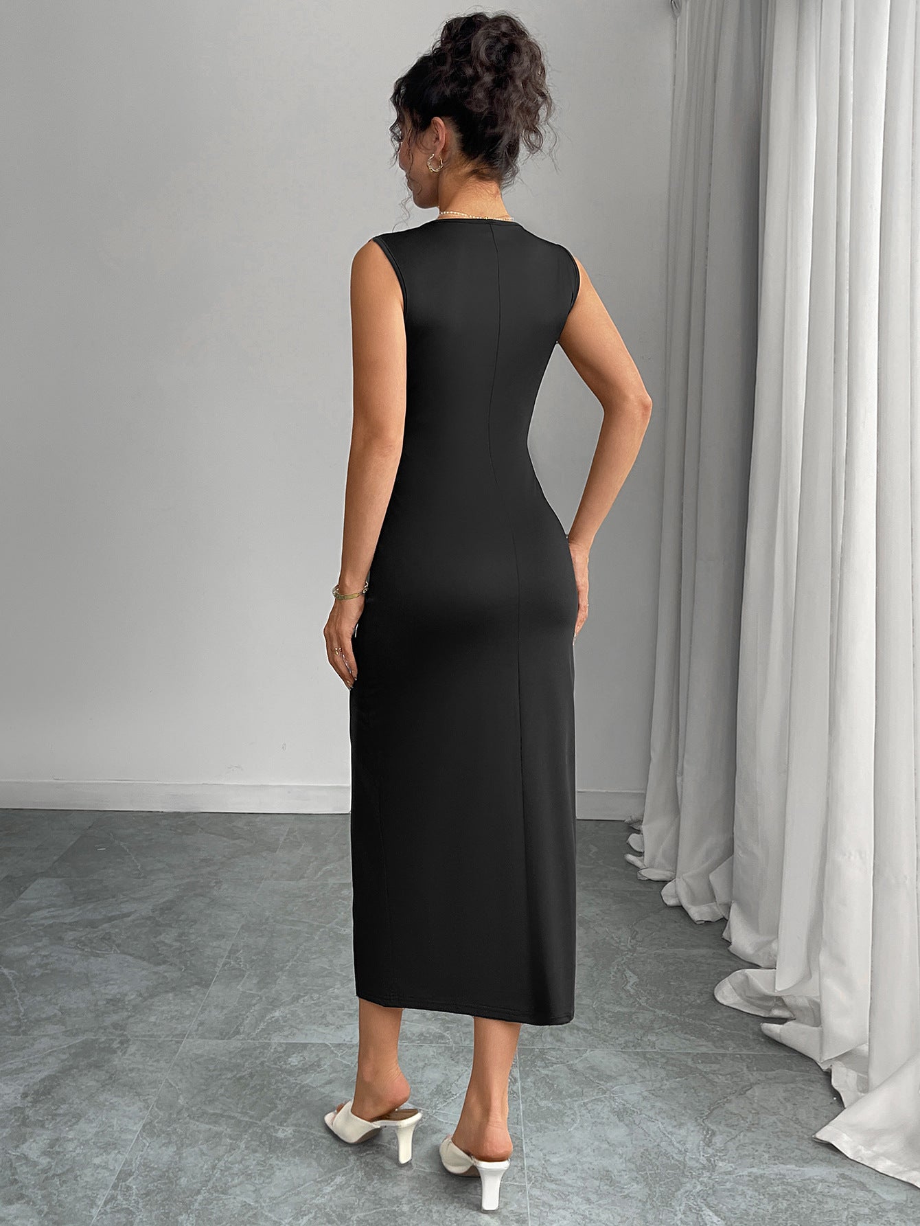 Back view of woman wearing a black sleeveless slit dress standing in a room with gray walls and white curtains.