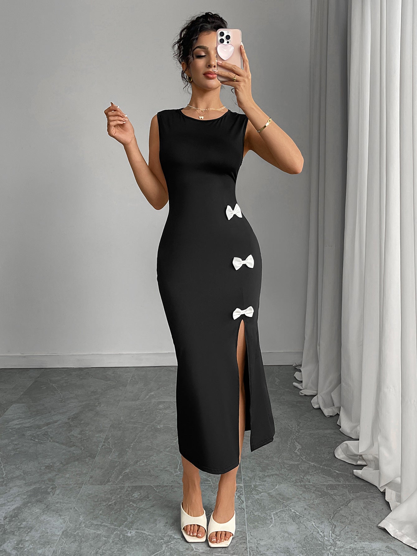 Woman taking a mirror selfie wearing a black slit dress with white bows in a room with gray walls and white curtains.