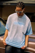 Man looks down while wearing blue quote tee, sunglasses, and chain necklace, standing beside a car.