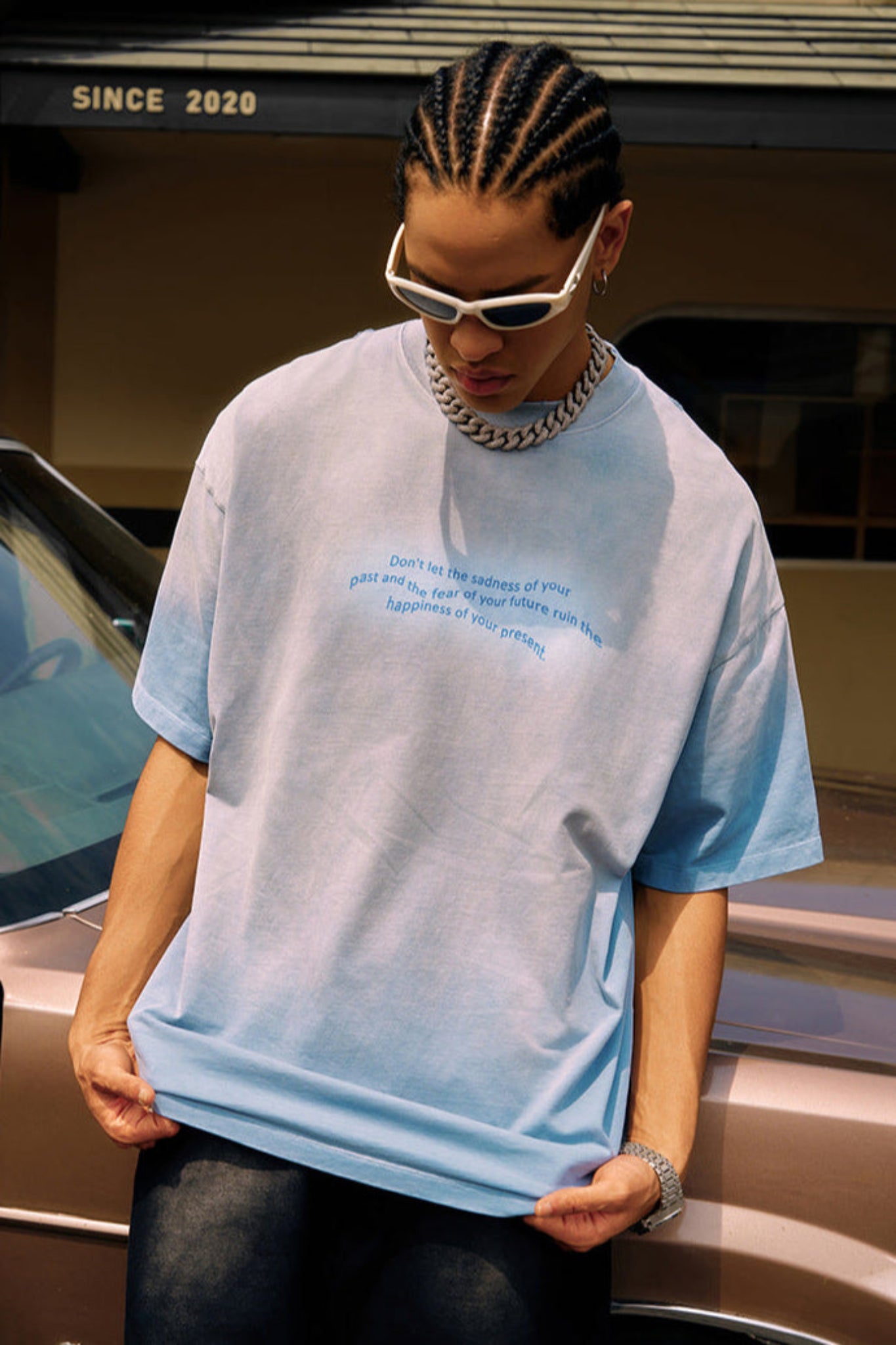 Man looks down while wearing blue quote tee, sunglasses, and chain necklace, standing beside a car.