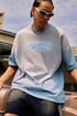Man in blue gradient tee and sunglasses sits on car hood, shot from a low angle.