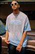 Man in blue oversized tee and accessories poses confidently in front of a car.

