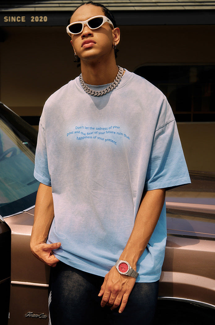 Man in blue oversized tee and accessories poses confidently in front of a car.

