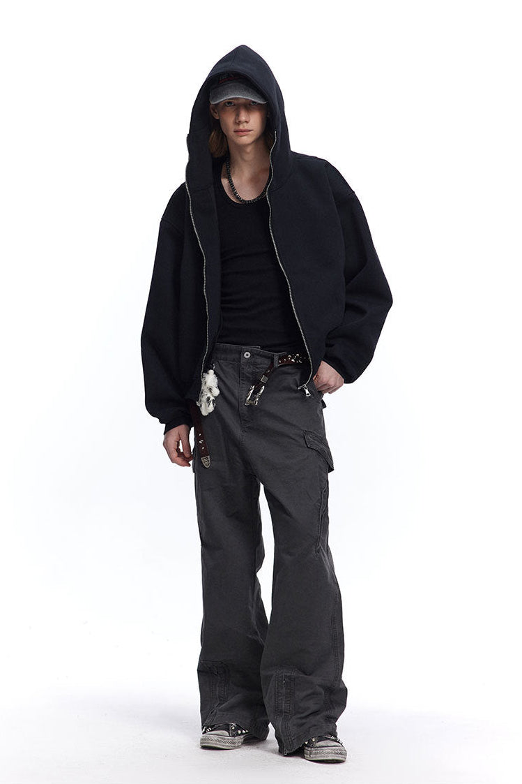 Full body view of a male model in a black boxy zip up hoodie, cap, black tank top, and wide-leg cargo pants.