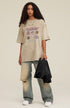Woman in oversized khaki pixel tee holding black shirt and wearing loose jeans