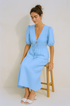 Woman wearing a light blue skirt set sitting on a wooden stool against a beige background