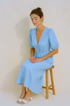 Woman wearing a light blue skirt set sitting on a wooden stool against a beige background