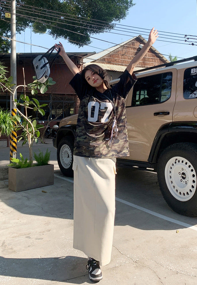 Model stands by SUV in cream cargo maxi skirt and camo jersey tee, arms raised.