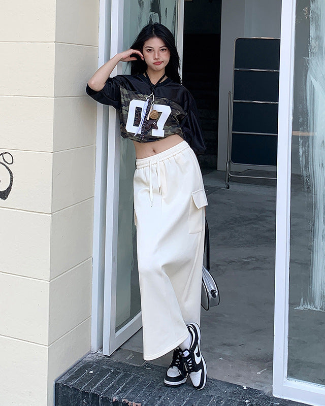 Model leans on doorway wearing cream maxi skirt with drawstring waist and cropped black top.