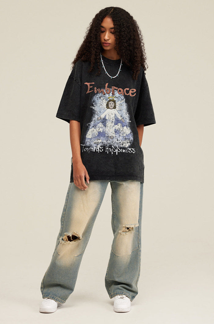 Model wearing black graphic T-shirt with angel design and distressed jeans, looking down with hands in pockets.