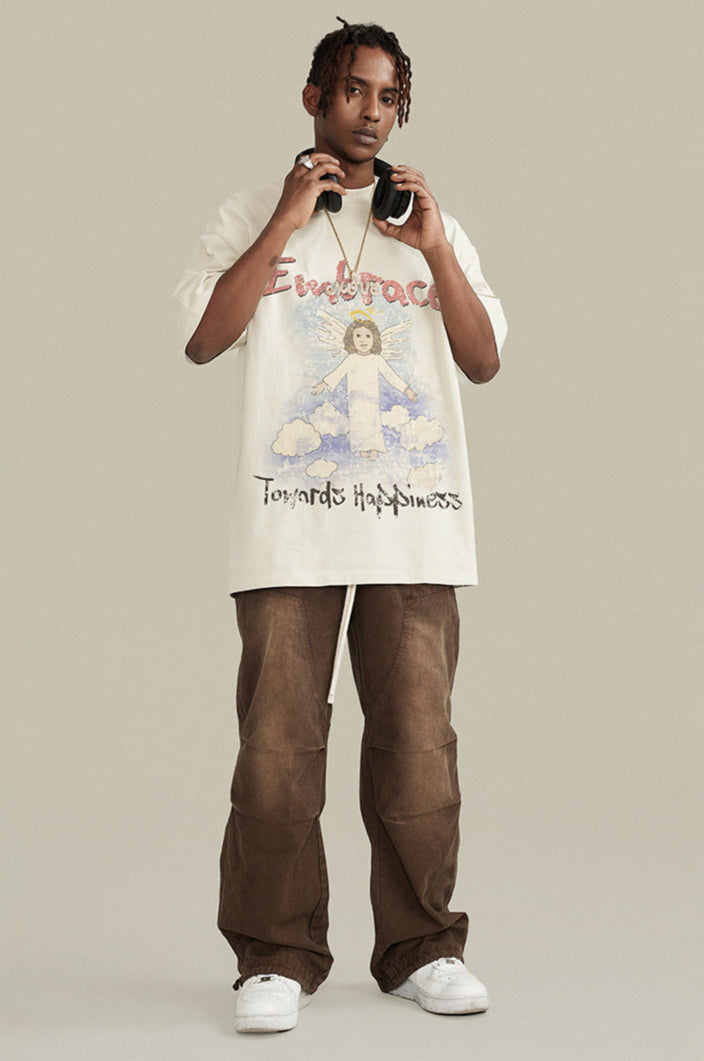 Model wearing cream angel graphic tee with headphones and brown cargo pants, standing pose.