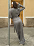 Back view of woman in a gray outfit standing against a building with a pole.