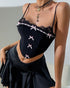 Woman leaning against a wall wearing a black lace-trimmed corset with pink bows and a black flowy mini skirt.