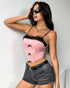 Woman posing in sunglasses, wearing a pink lace-trimmed corset top with black bows and black denim shorts.