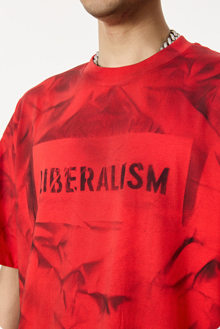 Close-up of red tee showing bold “LIBERALISM” text and box print over crumpled red texture.