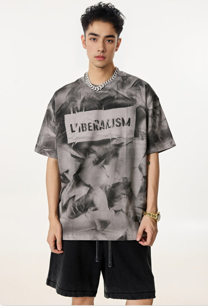 Man wears oversized gray graphic tee with “Liberalism” text and black shorts.
