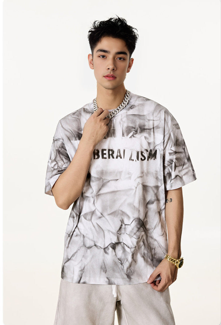 Man wears white “Liberalism” tee, hand resting on chain necklace.