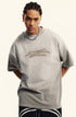 Man in light grey tee leans slightly forward with hands behind back, wearing chain necklace.