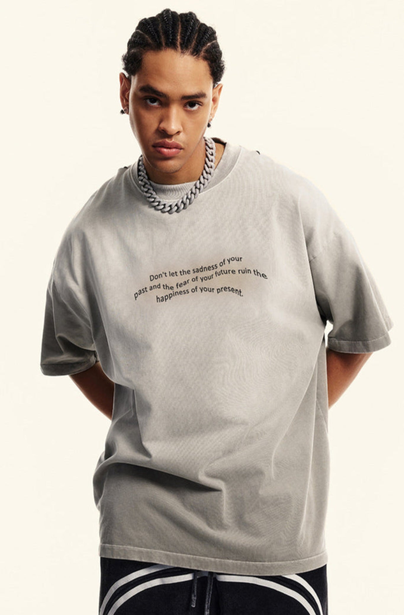 Man in light grey tee leans slightly forward with hands behind back, wearing chain necklace.