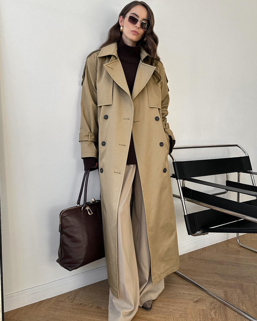 Model wearing a classic long beige double-breasted trench coat layered over a dark turtleneck and wide-leg trousers, accessorized with a dark brown leather bag and sunglasses.