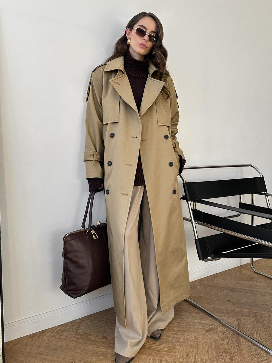 Model wearing a classic long beige double-breasted trench coat layered over a dark turtleneck and wide-leg trousers, accessorized with a dark brown leather bag and sunglasses.