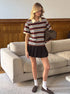 Model stands in living room wearing burgundy striped polo shirt with pleated black mini skirt and loafers.