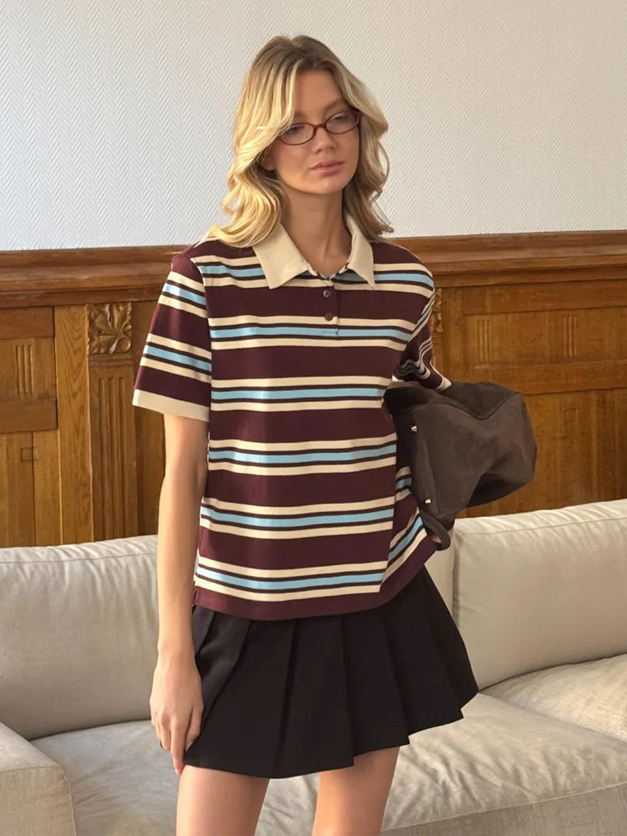 Model holds jacket while standing in burgundy striped polo shirt and pleated black skirt.