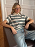 Model leans back in armchair wearing green striped polo shirt with relaxed light denim pants.