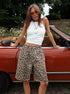 Woman wearing white sleeveless top and beige loose leopard print shorts, posing by a vintage red convertible.