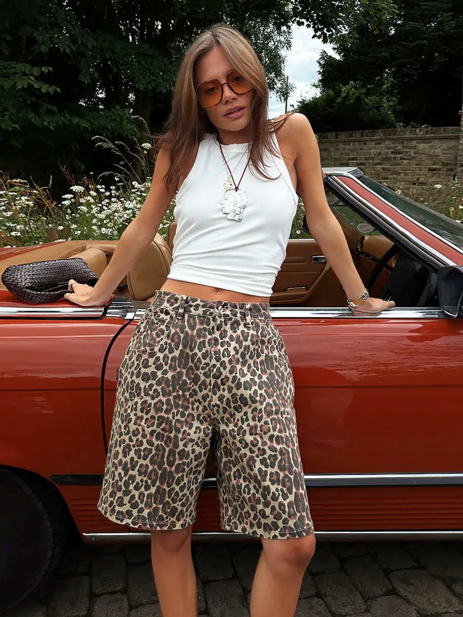 Woman wearing white sleeveless top and beige loose leopard print shorts, posing by a vintage red convertible.