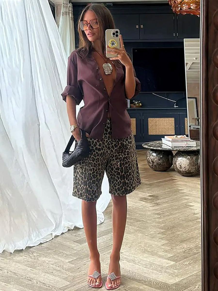 Woman taking mirror selfie in open brown shirt and loose leopard print shorts, accessorized with a black bag and heeled sandals.
