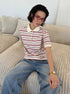 Model sits on beige sofa wearing cream and maroon striped polo shirt with red glasses.