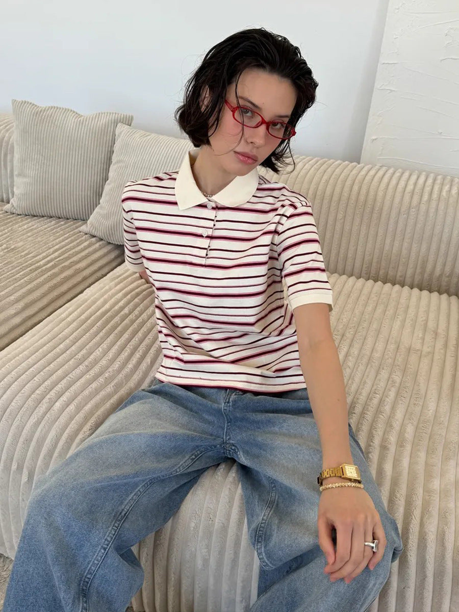 Model sits on beige sofa wearing cream and maroon striped polo shirt with red glasses.