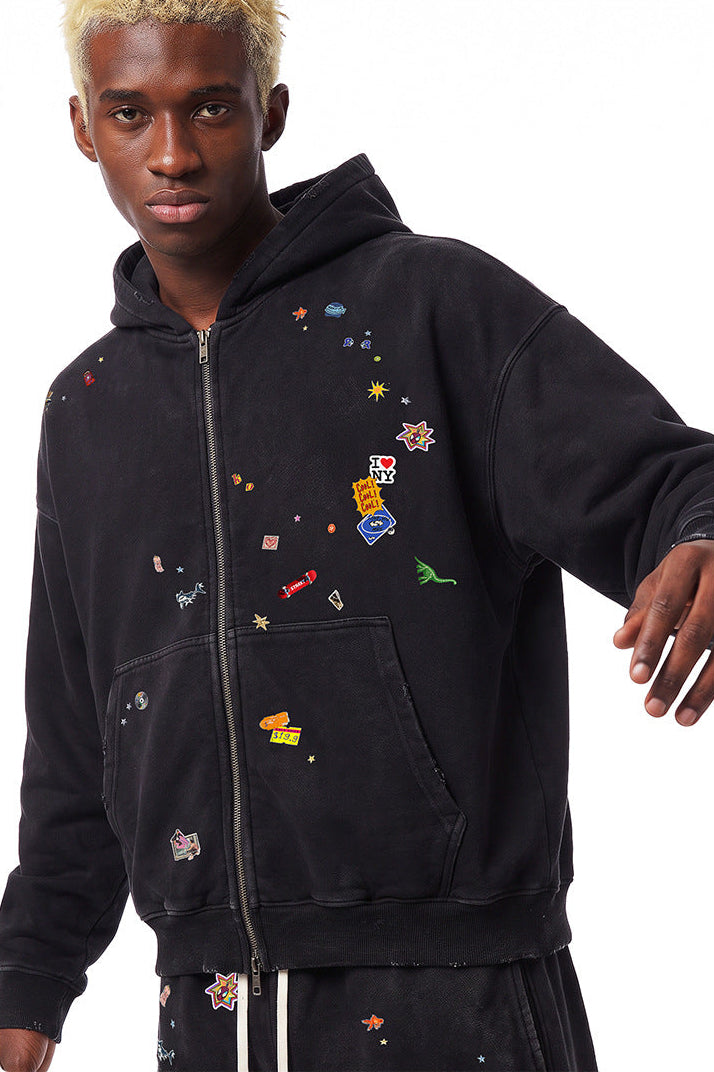 Medium shot of model styling a black boxy fit zip up hoodie with retro graphic patches and drop shoulder sleeves.