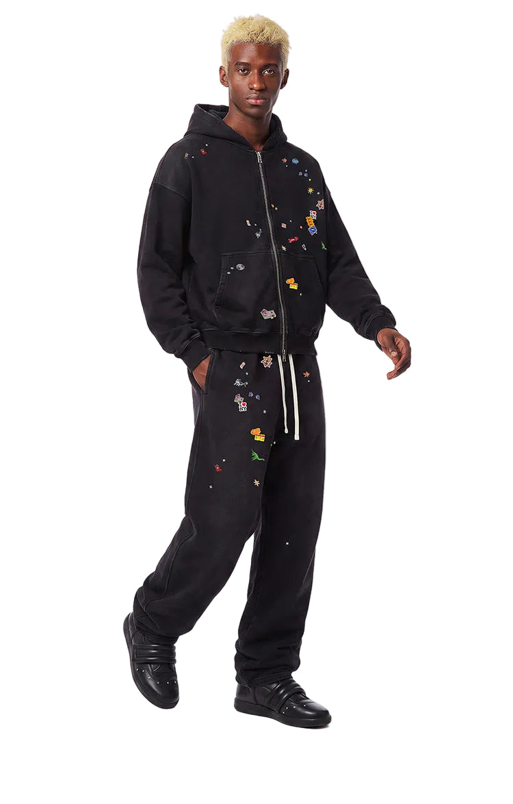 Side view of model walking in a black vintage washed zip up hoodie with graphic patches and loose sweatpants.