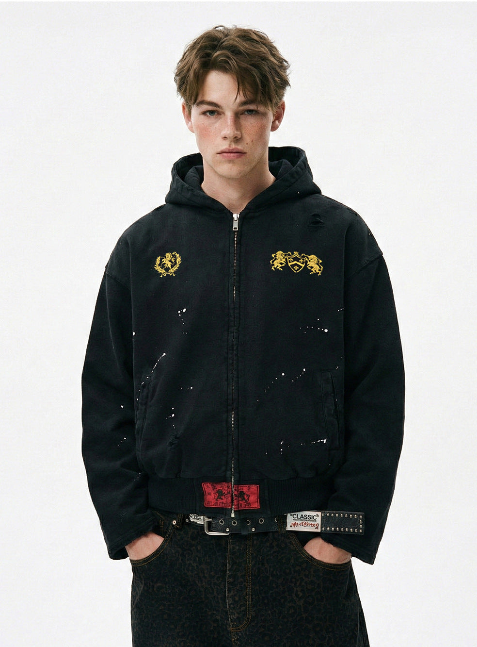 Model wearing a washed black distressed zip up hoodie with paint splatter and gold chest crest embroidery.
