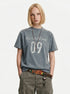 Blue-gray varsity graphic T-shirt styled with olive cargo pants and a leather belt.