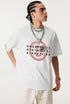 Man in white graphic tee glancing sideways with hands in pockets.