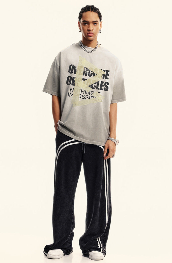 Model wears light gray oversized T-shirt with motivational text and taped arrow print, styled with black track pants.