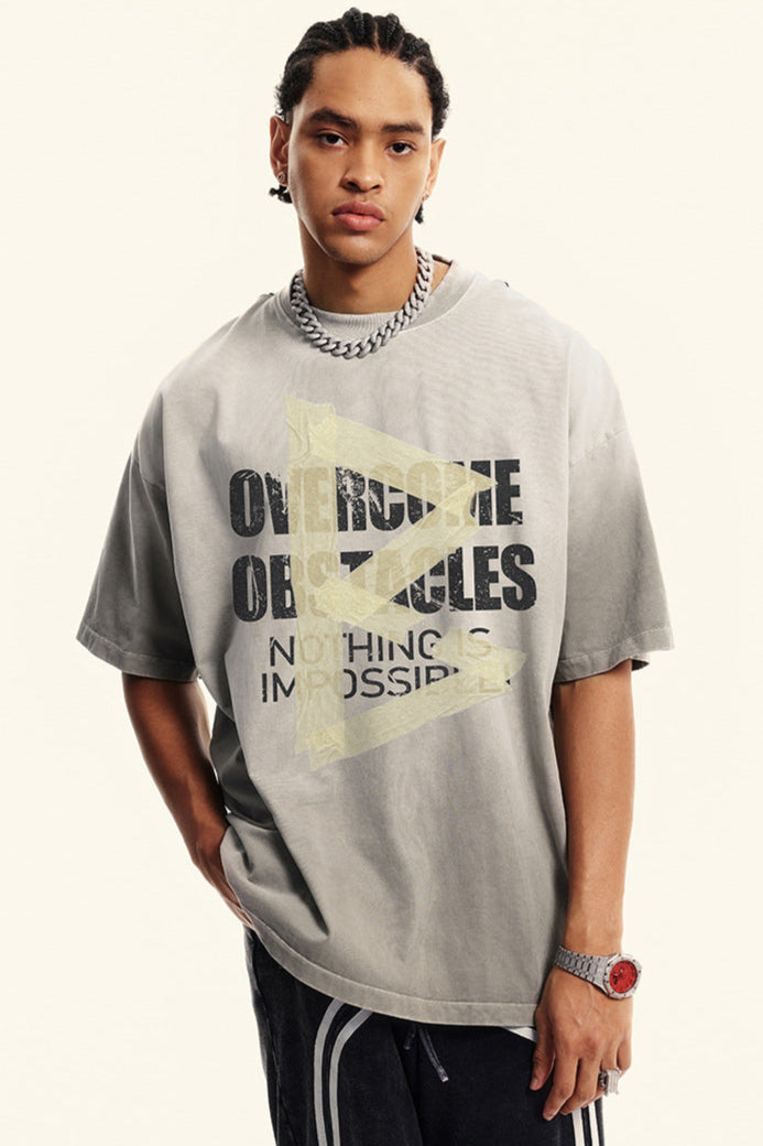 Light gray tee with “Overcome Obstacles” message and faded yellow arrow, styled in a streetwear look.