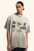 Model looks sideways wearing grey T-shirt with cracked type and yellow arrow print.