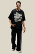 Man wearing black oversized T-shirt with motivational text graphic and striped track pants, standing on pavement near scaffolding.