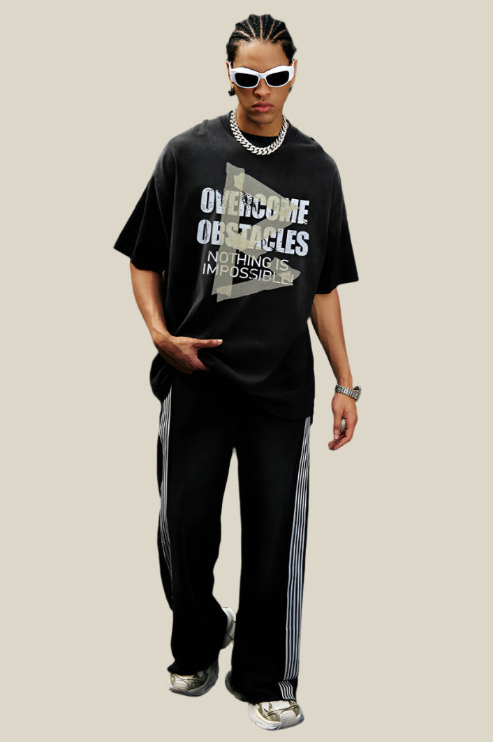 Man wearing black oversized T-shirt with motivational text graphic and striped track pants, standing on pavement near scaffolding.